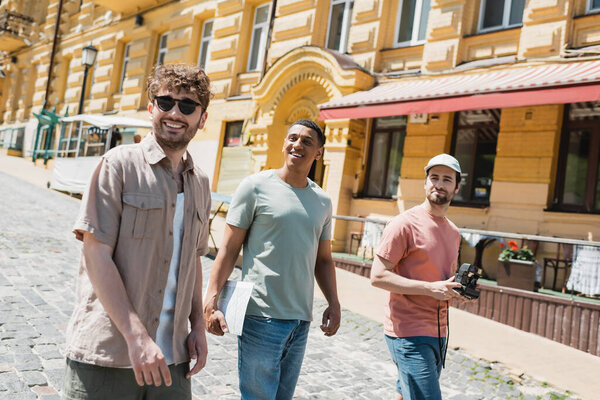 гид в солнцезащитных очках для мультикультурных туристов, прогуливающихся возле исторических зданий на Андреевском спуске в Киеве