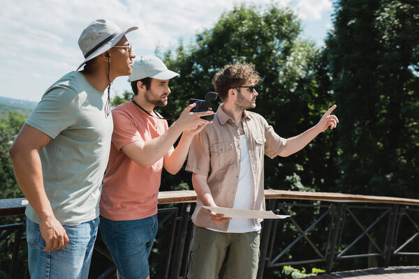 bearded man in sun hat taking photo on vintage camera near tour guide looking away and pointing with finger in city park