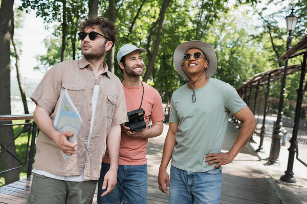 carefree african american tourist in sunglasses and sun hat looking away near guide and friend with vintage camera in city park