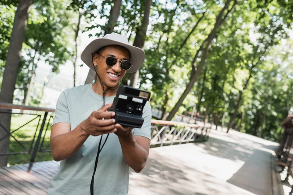戴着太阳帽和太阳镜的快乐的非洲游客在城市公园的人行道上用老式相机拍照 — 图库照片