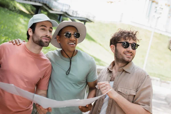 stock image cheerful interracial travelers in sun hats holding travel map and looking away on urban street