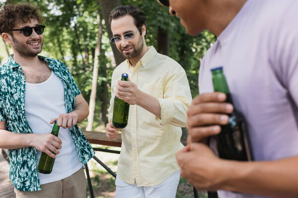 модный мужчина в стильных солнцезащитных очках держит бутылку свежего пива рядом с межрасовыми друзьями в летнем парке