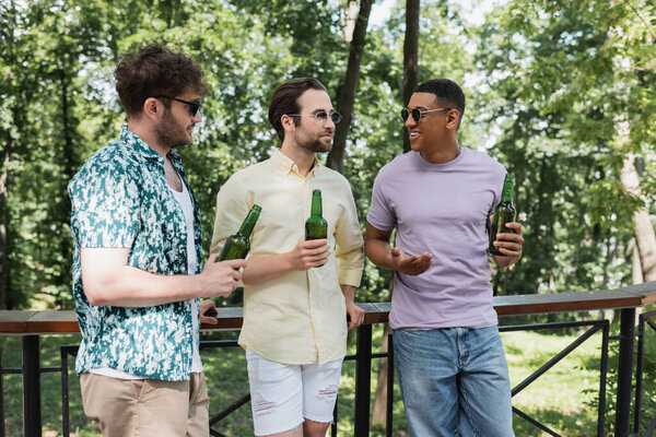 счастливые и стильные многонациональные друзья в солнечных очках, держащие свежее пиво и разговаривающие возле забора в городском парке