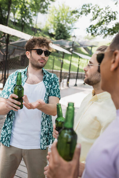 молодой и модный мужчина в солнечных очках, указывающий на бутылку пива рядом с размытыми межрасовыми друзьями в городском парке