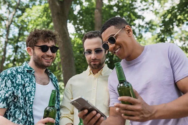 Улыбающийся Африканский Американец Показывает Мобильный Телефон Модным Друзьям Пивными Бутылками — стоковое фото