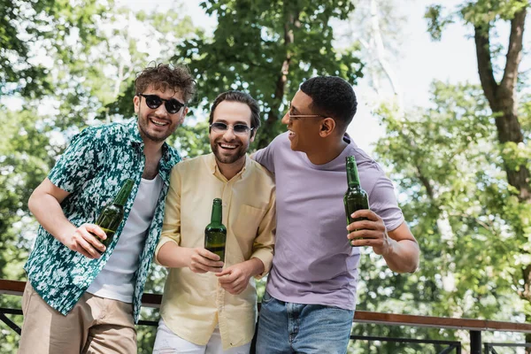 戴着太阳镜 拿着啤酒 在绿地公园里大笑的快乐而时髦的跨种族朋友 — 图库照片