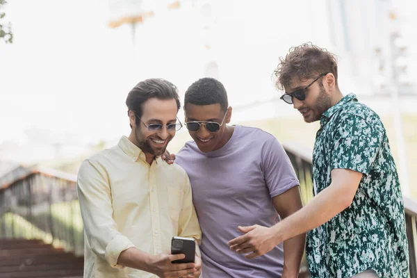 stock image Young man in sunglasses talking to interracial friends with smartphone on stairs in park 