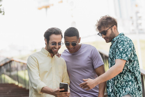 Молодой человек в солнечных очках разговаривает с межрасовыми друзьями со смартфоном на лестнице в парке 