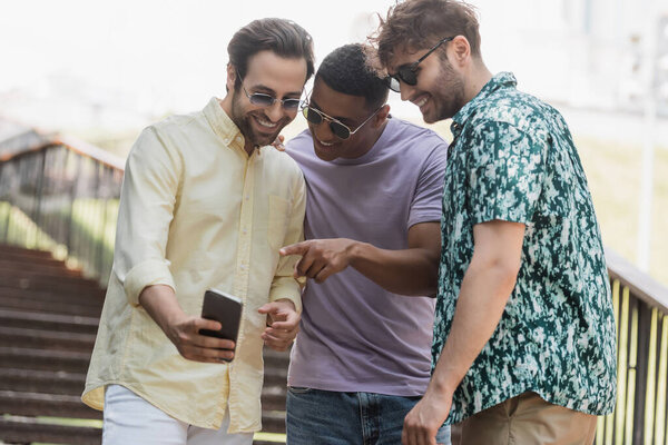Африканский американец указывает на смартфон рядом с веселыми друзьями в солнечных очках на лестнице в парке 
