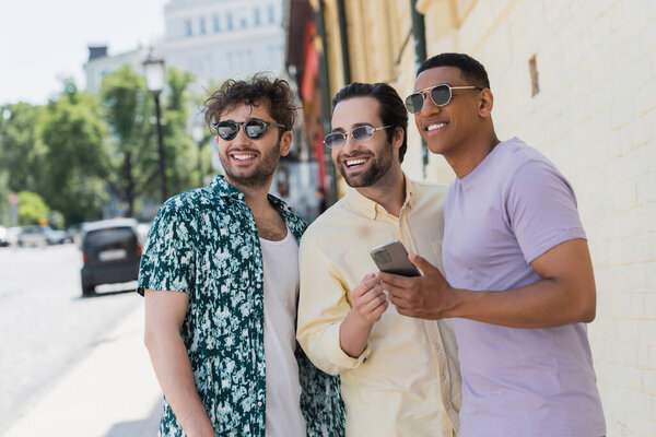 Улыбающиеся мультиэтнические мужчины в солнцезащитных очках, держащие мобильный телефон и смотрящие на улицу Киева 