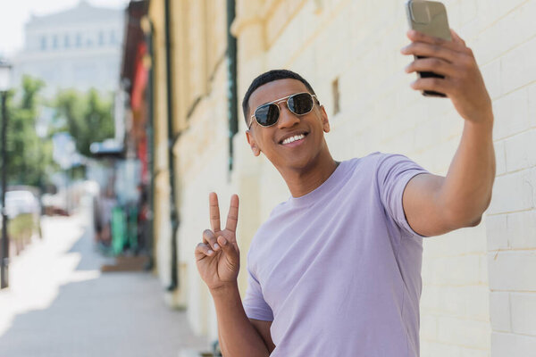 Smiling african american man in sunglasses showing peace sign while taking selfie on blurred urban street 