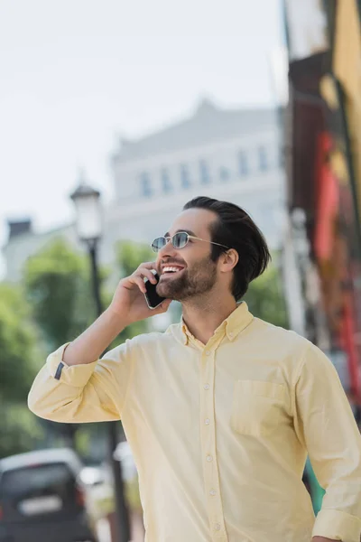 在城市街道上 戴着太阳镜 身穿衬衫 拿着智能手机说话的积极男子 — 图库照片