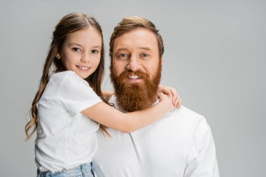 Gülümseyen çocuk sakallı babaya sarılıyor ve gri renkli kameraya bakıyor.  