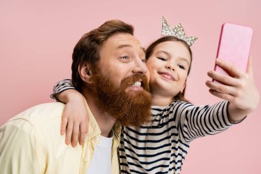 Tacından saç bandı takmış gülümseyen bir kız. Sakallı babasına sarılıyor. Pembe bir selfie çekerken.  