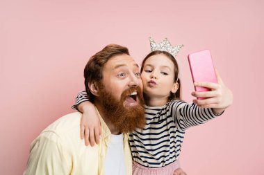 Taçlı kafa bandı takan, dudaklarını büzen ve sakallı babasıyla pembe bir selfie çeken küçük kız.  