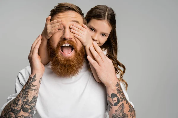 Preteen girl covering eyes of tattooed father with opened mouth isolated on grey