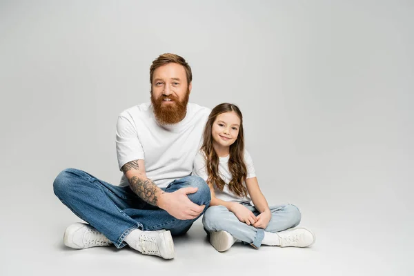 Carefree father and daughter in jeans and t-shirts sitting on grey background