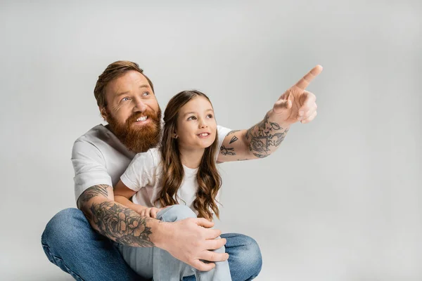 Cheerful tattooed man pointing with finger near preteen daughter isolated on grey