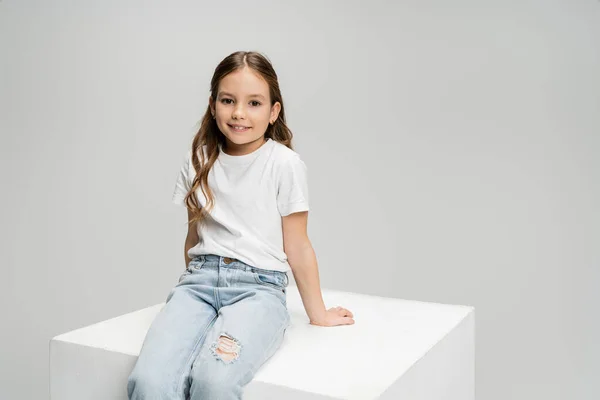 Smiling preteen girl in t-shirt and jeans sitting on cube isolated on grey