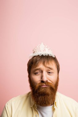 Taçlı sakallı üzgün bir adam pembe saç bandı takıyor.  
