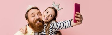 Pembe pankartta izole edilmiş selfie çekerken babasına taç bandıyla sarılan pozitif çocuk. 