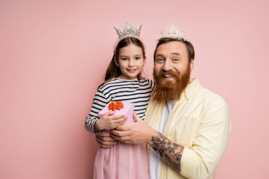 Taçlı dövmeli adam pembe arka planda küçük kızı yanında hediye tutuyor. 