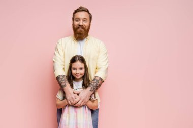 Neşeli bir adam reşit olmayan çocuğa sarılıyor ve pembe kameraya bakıyor.  