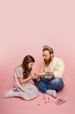 Sakallı babasının eline oje sürüp pembe arka planda saç bandı takan küçük bir kız. 