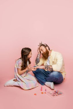 Preteen girl holding nail polish near hand of tired father with crown headband on pink background  clipart