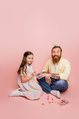 Pembe arka planda süslü makyaj malzemesi ve saç bandıyla babasının yanında oje tutan neşeli bir kız. 