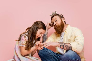 Pembe arka planda taç takan dövmeli babasının eline oje süren küçük bir kız. 