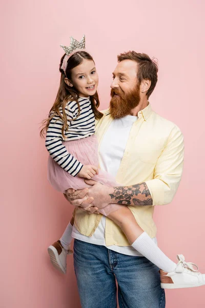 Tattooed man holding daughter with crown headband on pink background