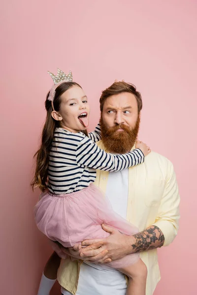Tattooed father holding preteen daughter in crown headband sticking out tongue on pink background