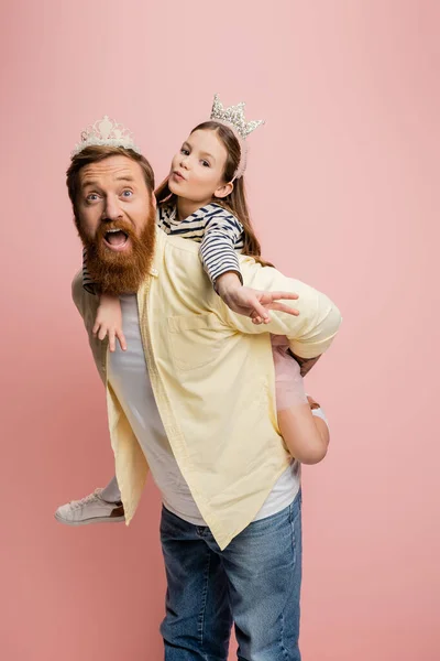 Preteen girl showing peace gesture while piggybacking on father isolated on pink