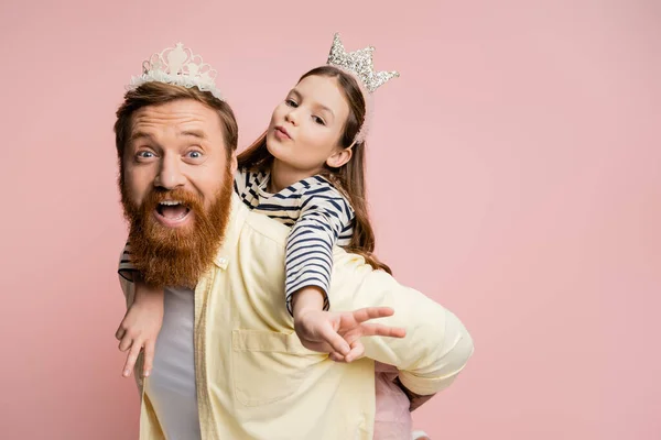 Preteen kid in crown headband showing peace sign while piggybacking on dad isolated on pink