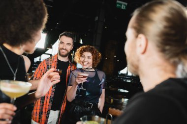 Smiling people holding cocktails near blurred interracial friends in bar at night  clipart