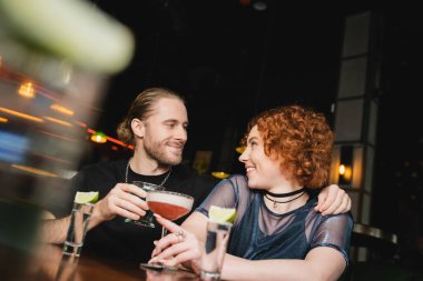 Positive man hugging red haired friend with cocktail near stand in bar  clipart