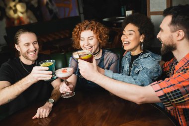 Cheerful multiethnic people clinking different cocktails in bar at night  clipart