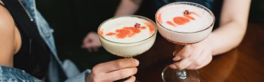 Cropped view of multiethnic girlfriends holding cold foam cocktails in bar in evening, banner  clipart