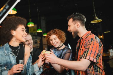 Cheerful friends holding lime and tequila near african american friend in bar  clipart