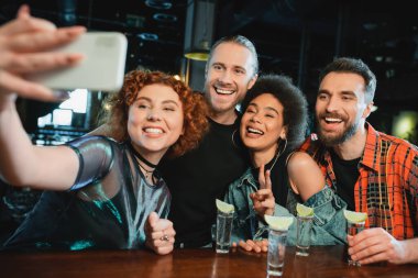 Gülümseyen çok ırklı arkadaşlar barda tekila yakınında akıllı telefondan selfie çekiyorlar. 