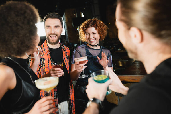 Positive interracial people holding glasses with cocktails and talking with friends in bar 