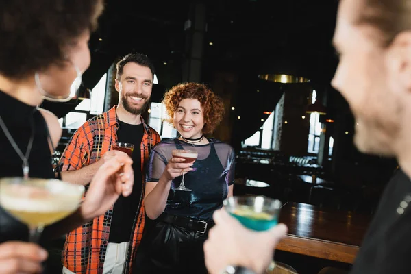 Cheerful people with cocktails meeting blurred interracial friends in bar