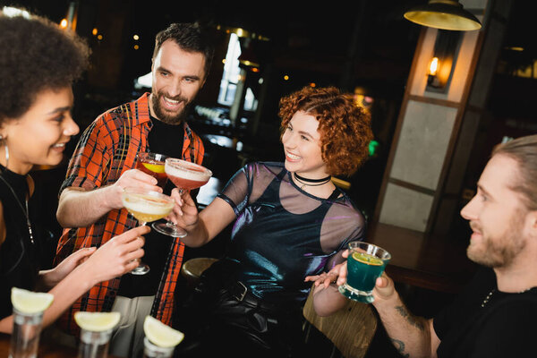 Overjoyed multiethnic friends holding glasses with cocktails near blurred tequila shots in bar 