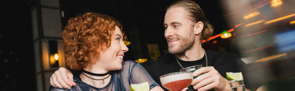 Smiling bearded man hugging redhead friend with cocktail in bar, banner 