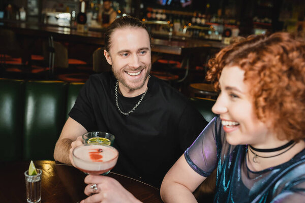 Cheerful man holding blue lagoon cocktail near blurred redhead friend in bar 