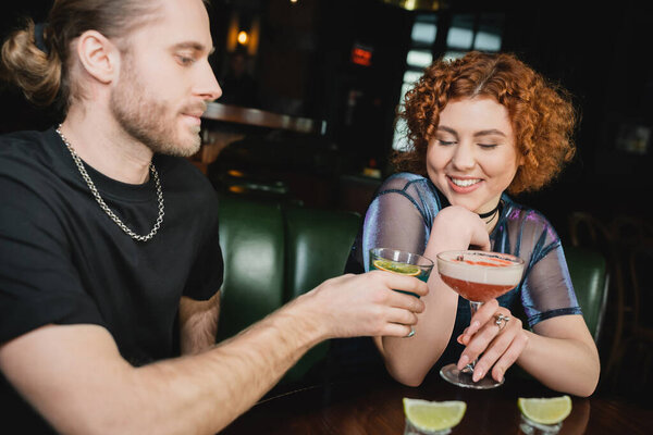 Positive red haired woman clinking cocktail with blurred friend in bar in evening