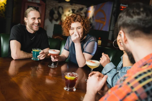 Cheerful redhead woman with cocktail sitting near multiethnic friends having fun in bar
