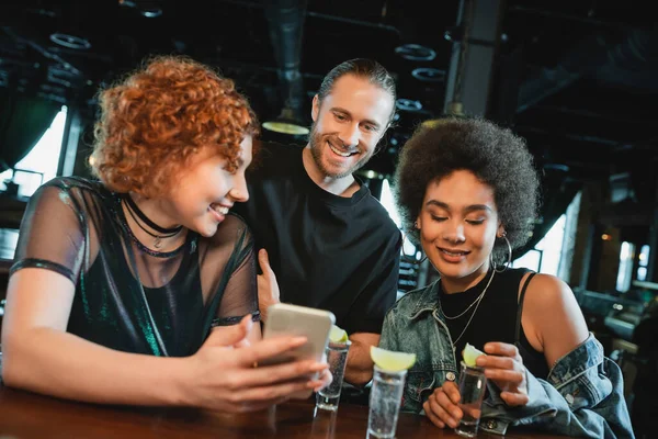 Smiling redhead woman holding smartphone near multiethnic friends and tequila in bar