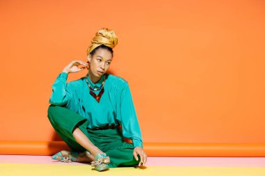 Renkli arka planda oturan parlak bluzlu ve pantolonlu modacı Afro-Amerikan modeli. 